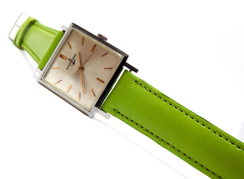 Pu&ograve; includere: Un orologio da polso quadrato con cassa argentata e quadrante chiaro. L'orologio ha lancette e indici color oro. &Egrave; dotato di un cinturino in pelle verde brillante. L'orologio &egrave; angolato su uno sfondo bianco.