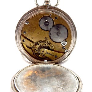 May include: Open silver pocket watch with visible gears and a gold-toned mechanism. The watch face is open, revealing the intricate inner workings. The watch has a crown and a hinged cover, and is set against a white background.