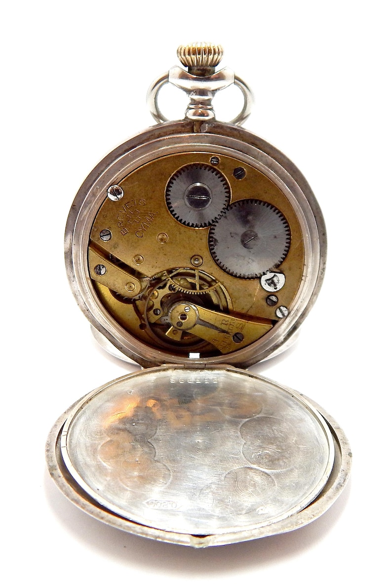 May include: Open silver pocket watch with visible gears and a gold-toned mechanism. The watch face is open, revealing the intricate inner workings. The watch has a crown and a hinged cover, and is set against a white background.