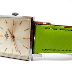 Pu&ograve; includere: Un orologio da polso vintage Ulysse Nardin con cassa quadrata color argento e quadrante chiaro con lancette e indici dorati. L'orologio ha un cinturino in pelle verde brillante. Il quadrante mostra il marchio Ulysse Nardin.
