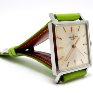 Pu&ograve; includere: Un orologio vintage con quadrante quadrato, cassa argentata e quadrante chiaro. L'orologio presenta lancette e indici dorati, ed &egrave; visibile il testo "ULYSSE NARDIN". Il cinturino &egrave; in pelle verde con retro marrone.
