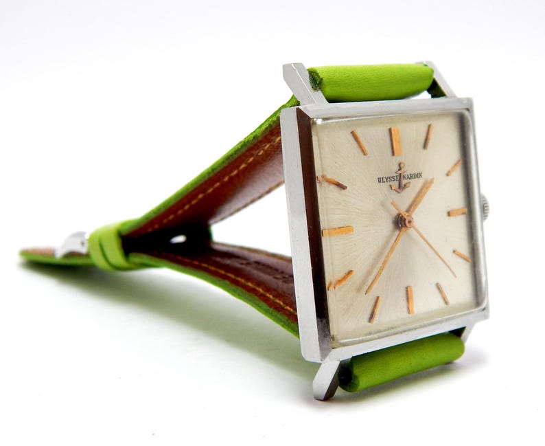 Pu&ograve; includere: Un orologio vintage con quadrante quadrato, cassa argentata e quadrante chiaro. L'orologio presenta lancette e indici dorati, ed &egrave; visibile il testo "ULYSSE NARDIN". Il cinturino &egrave; in pelle verde con retro marrone.