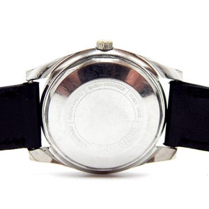 Puede incluir: Primer plano de un reloj de pulsera vintage, que muestra la parte trasera del reloj y su correa de cuero negro. El reloj tiene una caja plateada y una peque&ntilde;a corona. La parte trasera del reloj tiene el texto "SHOCK-ABSORBER" y "SWISS MADE".