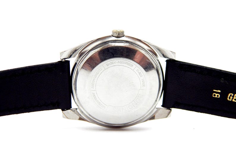 Puede incluir: Primer plano de un reloj de pulsera vintage, que muestra la parte trasera del reloj y su correa de cuero negro. El reloj tiene una caja plateada y una peque&ntilde;a corona. La parte trasera del reloj tiene el texto "SHOCK-ABSORBER" y "SWISS MADE".