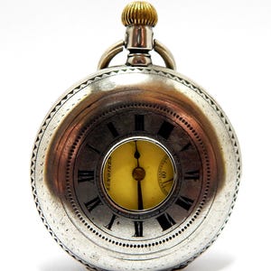 May include: A silver-toned pocket watch with a round case and a textured edge. The face features a yellow center dial with black hands, surrounded by a metallic ring with black Roman numerals and the text Longines. A gold-toned crown sits on top.