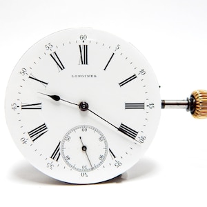 May include: A white Longines watch face with black Roman numerals and hands. A smaller seconds dial is at the bottom. The winding stem and crown are gold-colored. The watch face is set against a white background.