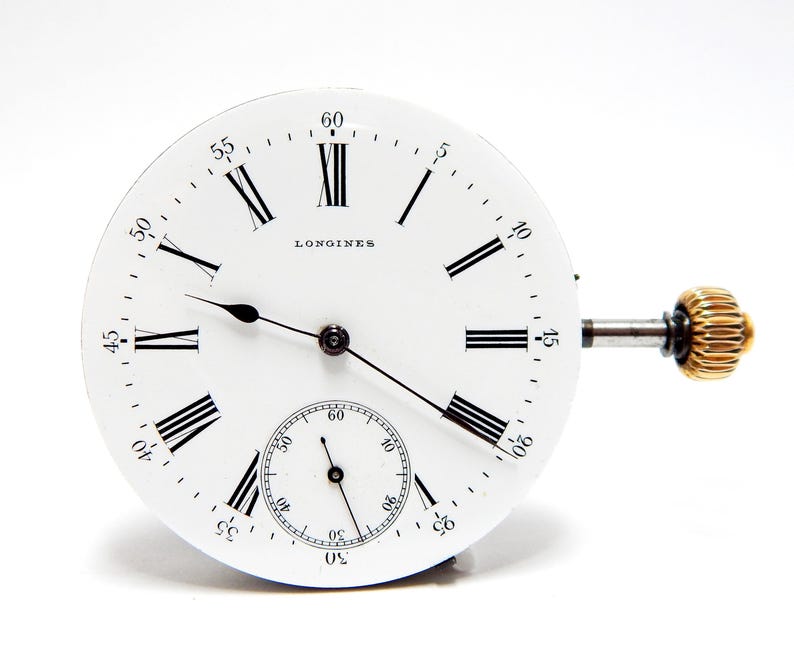 May include: A white Longines watch face with black Roman numerals and hands. A smaller seconds dial is at the bottom. The winding stem and crown are gold-colored. The watch face is set against a white background.