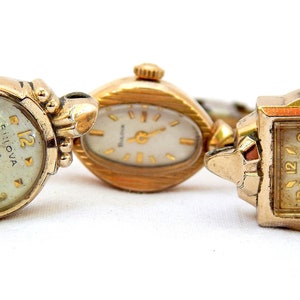 May include: Three gold-toned wristwatches with different styles of faces and bands. The watches are arranged in a row on a white surface.