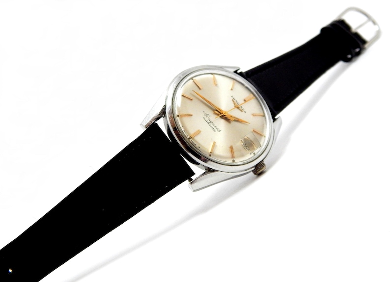Puede incluir: Un reloj de pulsera vintage con caja plateada, esfera clara y agujas e indicadores dorados. El reloj tiene una correa de cuero negro. La esfera del reloj muestra el texto "Longines Conquest Automatic".