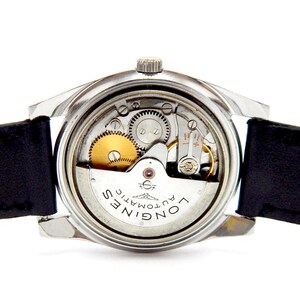 Puede incluir: Primer plano de un reloj autom&aacute;tico Longines vintage, mostrando sus intrincados mecanismos internos. El reloj tiene una caja plateada, una correa de cuero negro y un sistema de engranajes visible con componentes dorados y plateados. La esfera del reloj muestra "Longines Automatic".