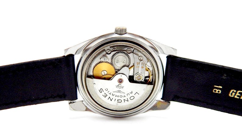 Puede incluir: Primer plano de un reloj autom&aacute;tico Longines vintage, mostrando sus intrincados mecanismos internos. El reloj tiene una caja plateada, una correa de cuero negro y un sistema de engranajes visible con componentes dorados y plateados. La esfera del reloj muestra "Longines Automatic".