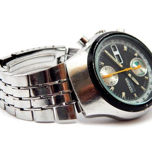 May include: A silver-toned Citizen automatic chronograph watch with a black face, featuring orange and white accents. The watch has a stainless steel band and is set against a white background.