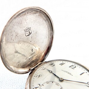 K&ouml;nnte beinhalten: Eine silberne Taschenuhr mit ge&ouml;ffnetem Deckel, die das Zifferblatt und die Innenseite des Deckels zeigt. Das Zifferblatt ist wei&szlig; mit schwarzen Ziffern und Zeigern. Die Innenseite des Deckels hat ein Logo und Text. Vintage-Taschenuhr.