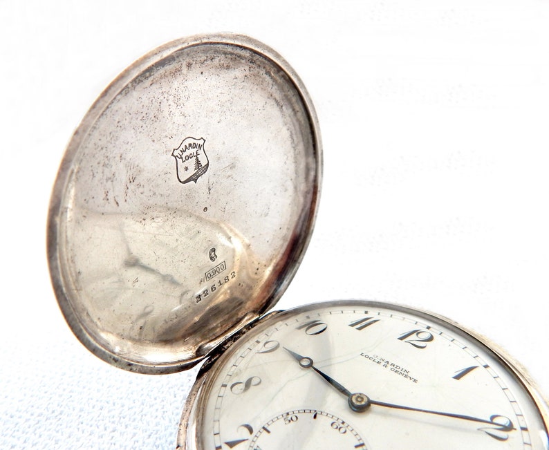 K&ouml;nnte beinhalten: Eine silberne Taschenuhr mit ge&ouml;ffnetem Deckel, die das Zifferblatt und die Innenseite des Deckels zeigt. Das Zifferblatt ist wei&szlig; mit schwarzen Ziffern und Zeigern. Die Innenseite des Deckels hat ein Logo und Text. Vintage-Taschenuhr.