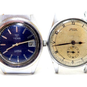 Puede incluir: Primer plano de dos relojes de pulsera vintage. El primer reloj tiene una esfera azul oscuro con la marca "YEMA" y el texto "17 JEWELS ANTICHOC". El segundo reloj tiene una esfera color crema con la marca "MENTOR" y un pequeño dial de segundos.