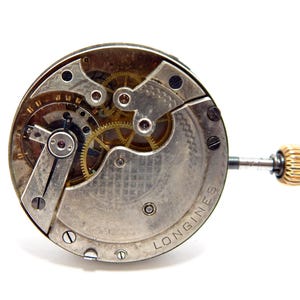 May include: Close-up of a vintage Longines watch movement. The mechanism features intricate gears, screws, and a gold-colored crown. The watch components are set against a silver-toned base with the brand name visible.