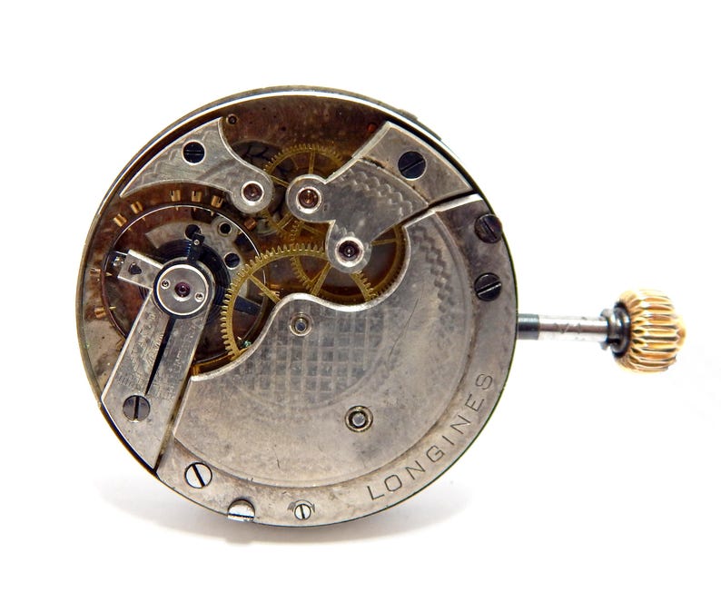 May include: Close-up of a vintage Longines watch movement. The mechanism features intricate gears, screws, and a gold-colored crown. The watch components are set against a silver-toned base with the brand name visible.