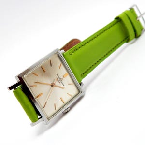 Pu&ograve; includere: Un orologio da polso quadrato vintage con cassa argentata e cinturino in pelle verde chiaro. Il quadrante dell'orologio &egrave; di colore champagne chiaro con lancette e indici dorati. Il cinturino &egrave; largo circa 2 cm.