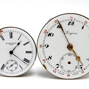 May include: Two antique watch faces. One is marked "Ulysse Nardin Locle" with Roman numerals and a subdial. The other is a "Longines" watch face with black numerals, red accents, and a subdial. Both have black hands.