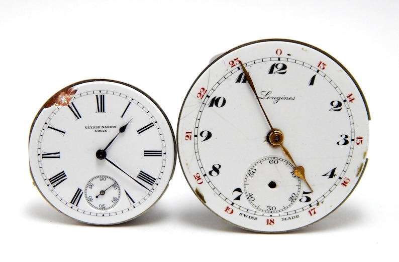May include: Two antique watch faces. One is marked "Ulysse Nardin Locle" with Roman numerals and a subdial. The other is a "Longines" watch face with black numerals, red accents, and a subdial. Both have black hands.