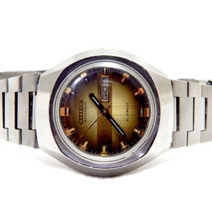 May include: A silver Citizen automatic wristwatch with a brown face and a silver metal band. The watch face has gold-colored hour markers and a date window that reads "01023".