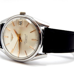Puede incluir: Un reloj de pulsera autom&aacute;tico Longines Conquest plateado con una correa de cuero negro. La esfera del reloj es de color crema con agujas e indicadores dorados. La fecha se muestra a las 3 en punto. El reloj est&aacute; en &aacute;ngulo, mostrando la correa y la esfera.