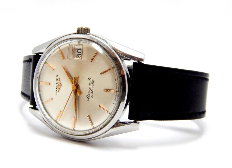 Puede incluir: Un reloj de pulsera autom&aacute;tico Longines Conquest plateado con una correa de cuero negro. La esfera del reloj es de color crema con agujas e indicadores dorados. La fecha se muestra a las 3 en punto. El reloj est&aacute; en &aacute;ngulo, mostrando la correa y la esfera.