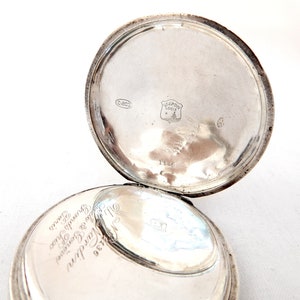 May include: Open silver pocket watch with the back lid ajar. The inside of the lid has the text "Ulysse Nardin Locle" and other markings. The watch is on a white surface.