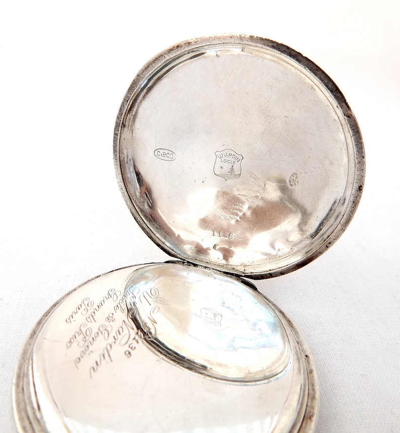 May include: Open silver pocket watch with the back lid ajar. The inside of the lid has the text "Ulysse Nardin Locle" and other markings. The watch is on a white surface.