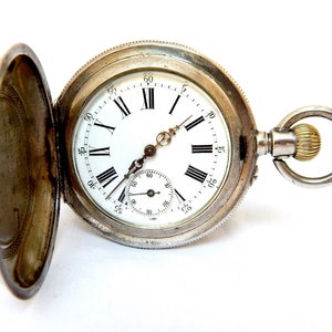 Puede incluir: Reloj de bolsillo plateado con tapa abierta, que revela una esfera blanca con números romanos negros y un pequeño dial de segundos. El reloj tiene una corona dorada y un lazo de cadena plateado.