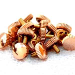 May include: A pile of small, copper-colored watch crowns with a ridged edge and a cylindrical post. The crowns are clustered together against a white background, showcasing their metallic sheen and intricate design. These watch parts are for watch repair or crafting.
