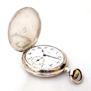 May include: A silver pocket watch with the cover open, revealing a white face with black numerals and hands. A smaller dial is at the bottom. The watch has a gold-colored ring for attaching a chain. The back of the watch cover has markings.