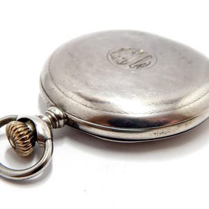 May include: A silver pocket watch with a textured face and a decorative ring. The watch has a circular design with an engraved emblem on the cover. The watch's ring is gold-colored and has a textured design.