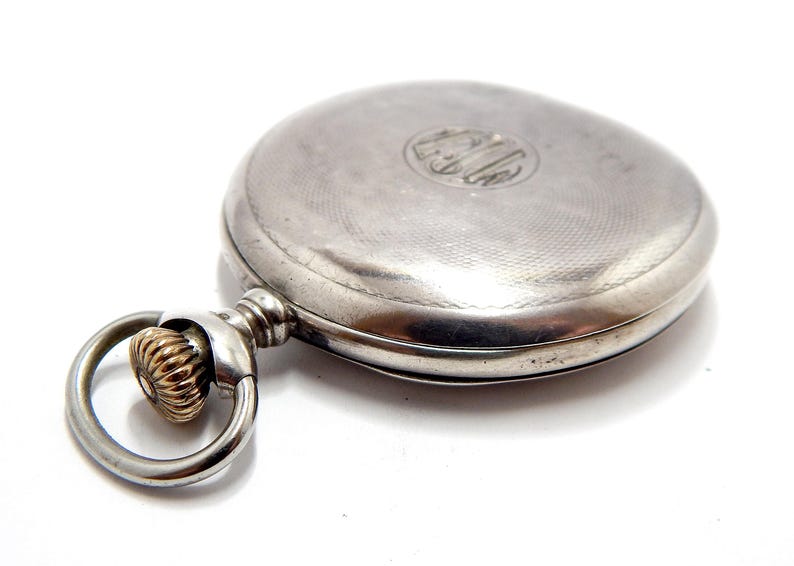 May include: A silver pocket watch with a textured face and a decorative ring. The watch has a circular design with an engraved emblem on the cover. The watch's ring is gold-colored and has a textured design.