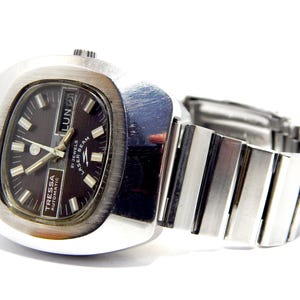 Könnte beinhalten: Eine Vintage Tressa Automatic Uhr mit silberfarbenem Gehäuse und Armband. Das Zifferblatt ist dunkel mit silbernen Zeigern und Markierungen und zeigt den Wochentag an. Das Armband besteht aus rechteckigen Metallgliedern.