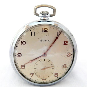 Op de afbeelding: Een zilveren zakhorloge met een witte wijzerplaat en zwarte Romeinse cijfers. Het horloge heeft een kleine secondewijzer-wijzerplaat en de tekst "CYMA" en "SWISS MADE" op de wijzerplaat.