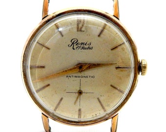 Peut inclure: Une montre-bracelet dorée avec un cadran blanc et des aiguilles noires. Le cadran de la montre porte le texte "Renis 17 Rubis" et "ANTIMAGNETIC".