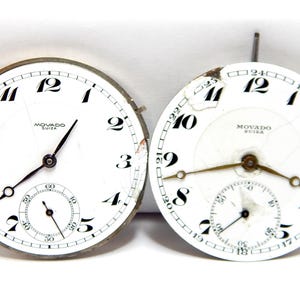 May include: Two vintage Movado watch faces with white dials, black numerals, and sub-dials. The faces show signs of age, with some wear and tear. The text "MOVADO SUISSA" is visible on both.