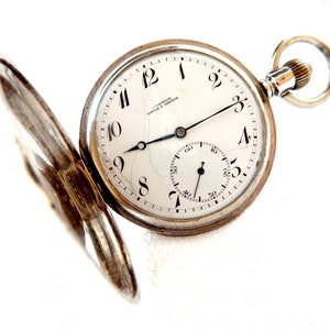 K&ouml;nnte beinhalten: Eine silberne Taschenuhr mit offenem Deckel, die ein wei&szlig;es Zifferblatt mit schwarzen Ziffern und Zeigern zeigt. Das Zifferblatt hat ein kleineres Hilfszifferblatt und die Marke "U. Nardin Locle & Geneve" ist aufgedruckt.