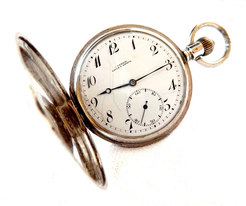 K&ouml;nnte beinhalten: Eine silberne Taschenuhr mit offenem Deckel, die ein wei&szlig;es Zifferblatt mit schwarzen Ziffern und Zeigern zeigt. Das Zifferblatt hat ein kleineres Hilfszifferblatt und die Marke "U. Nardin Locle & Geneve" ist aufgedruckt.