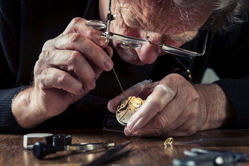May include: A close-up of a watchmaker at work, meticulously repairing a watch. The watchmaker is wearing a loupe and using small tools to work on the watch's intricate internal mechanisms. The watch parts are gold and silver.