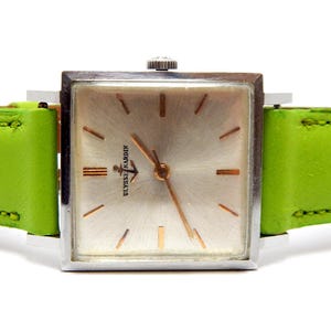 Pu&ograve; includere: Un orologio da polso vintage Ulysse Nardin con quadrante quadrato argentato e lancette e indici dorati. L'orologio ha un cinturino in pelle verde brillante. Il quadrante dell'orologio presenta il logo Ulysse Nardin.