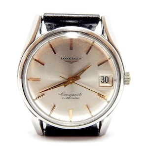 Puede incluir: Un reloj de pulsera autom&aacute;tico Longines Conquest plateado con una correa de cuero negro. La esfera del reloj es plateada con agujas e indicadores dorados, y una visualizaci&oacute;n de la fecha a las 3 en punto. El reloj tiene la palabra "Longines" en la parte superior y "Conquest automatic" en la inferior.