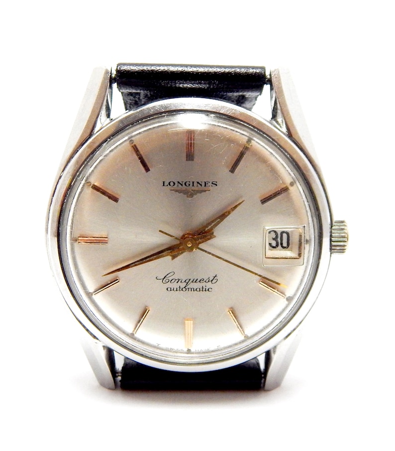 Puede incluir: Un reloj de pulsera autom&aacute;tico Longines Conquest plateado con una correa de cuero negro. La esfera del reloj es plateada con agujas e indicadores dorados, y una visualizaci&oacute;n de la fecha a las 3 en punto. El reloj tiene la palabra "Longines" en la parte superior y "Conquest automatic" en la inferior.
