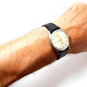 Puede incluir: Un reloj de pulsera vintage con caja plateada y esfera crema. El reloj tiene agujas e indicadores negros, y una correa de cuero negro. El reloj est&aacute; sujeto a una mu&ntilde;eca.