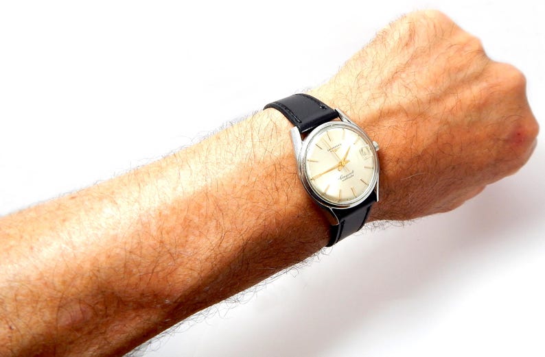 Puede incluir: Un reloj de pulsera vintage con caja plateada y esfera crema. El reloj tiene agujas e indicadores negros, y una correa de cuero negro. El reloj est&aacute; sujeto a una mu&ntilde;eca.