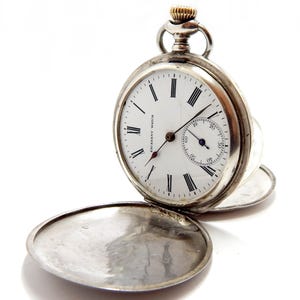 May include: A silver pocket watch with a white face and black Roman numerals. The watch has a small seconds dial and the cover is open. The watch is labeled "ESCASANY WATCH". The crown is gold-colored.