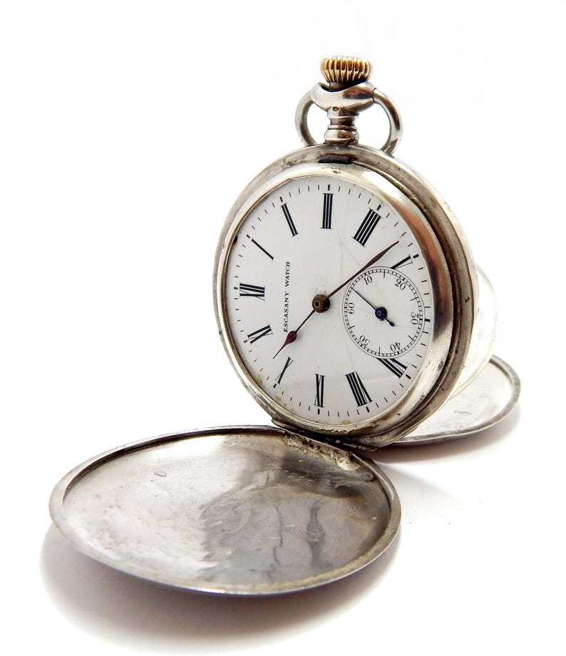May include: A silver pocket watch with a white face and black Roman numerals. The watch has a small seconds dial and the cover is open. The watch is labeled "ESCASANY WATCH". The crown is gold-colored.