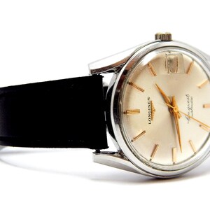Puede incluir: Un reloj Longines vintage con caja plateada y esfera crema. El reloj presenta agujas e indicadores dorados y una correa de cuero negro. El reloj est&aacute; sobre un fondo blanco.