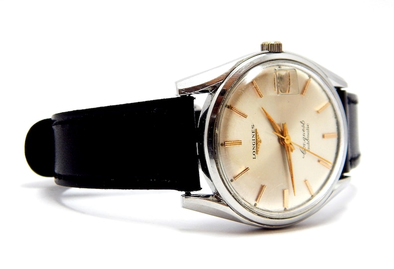 Puede incluir: Un reloj Longines vintage con caja plateada y esfera crema. El reloj presenta agujas e indicadores dorados y una correa de cuero negro. El reloj est&aacute; sobre un fondo blanco.